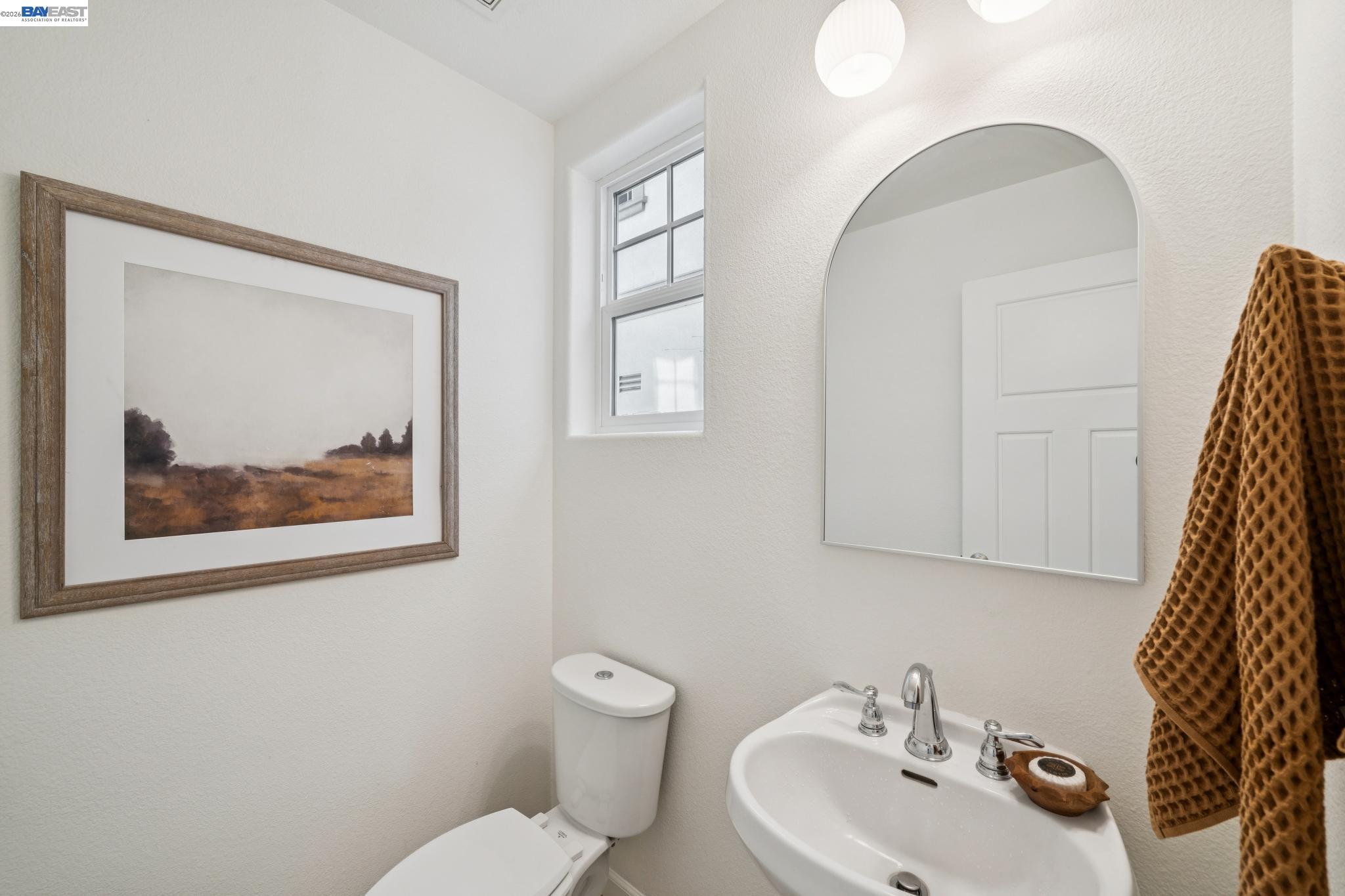 Bathroom with a pedestal sink, arched mirror, small window, and framed landscape print.