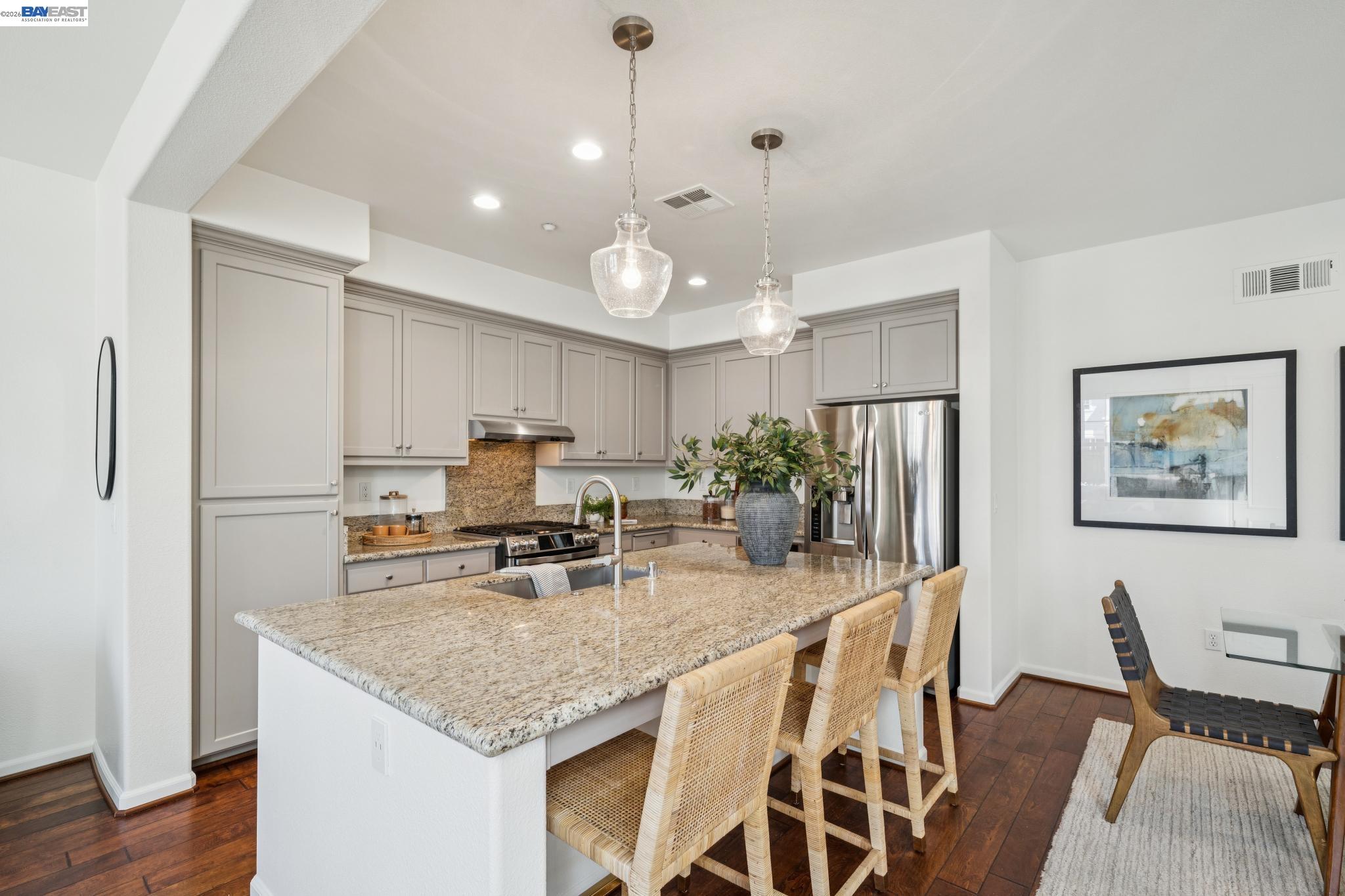 Kitchen with a granite island, woven bar stools, taupe cabinets, and stainless steel appliances.