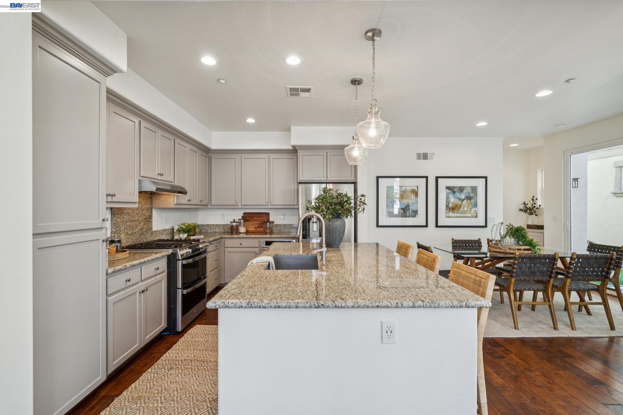 Kitchen with a large granite island, gray cabinets, stainless steel appliances, and an adjoining dining area.