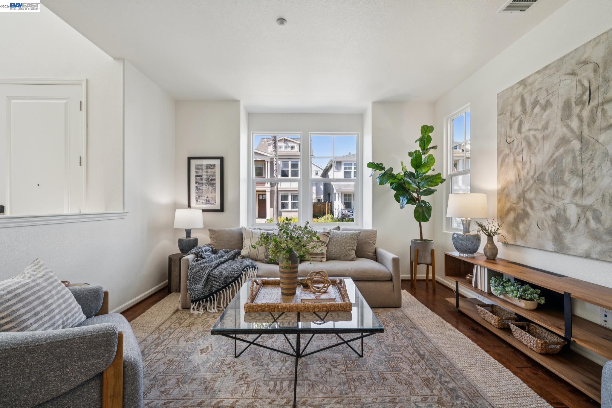 Living room with a beige sofa, glass coffee table, patterned rug, and large sunny windows.
