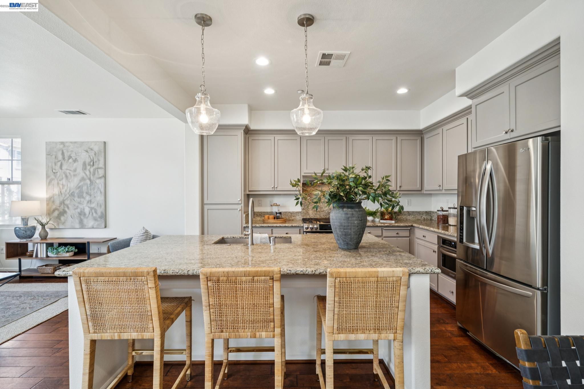Open-plan kitchen with a granite island, woven bar stools, taupe cabinets, and stainless steel appliances.