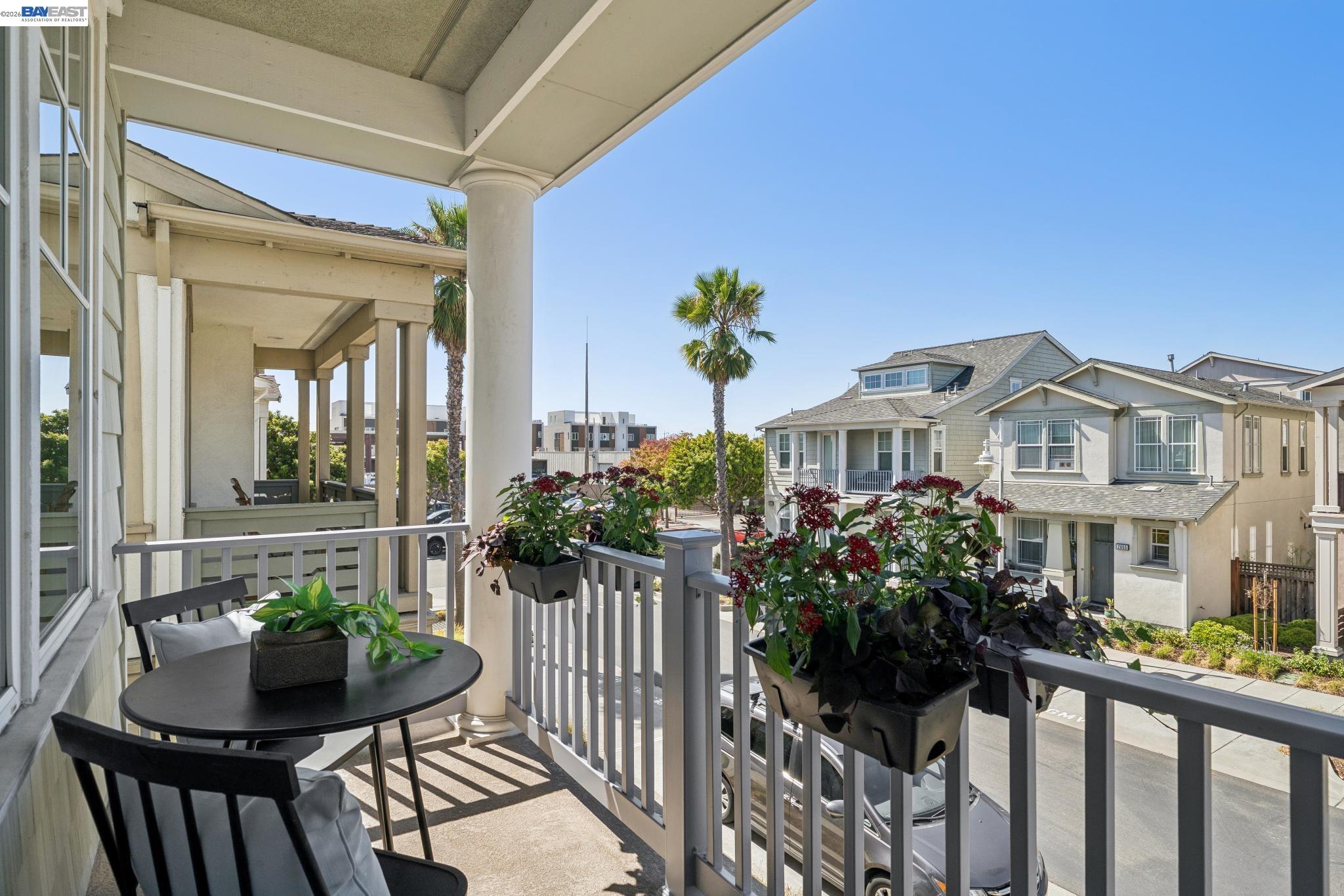 Covered balcony with patio seating and flower planters overlooking neighboring homes and a palm-lined street.