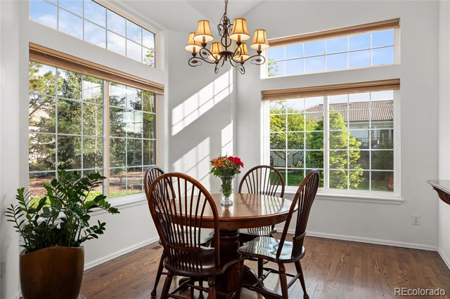 Dining room with a round wooden table, chandelier, and tall windows overlooking greenery.