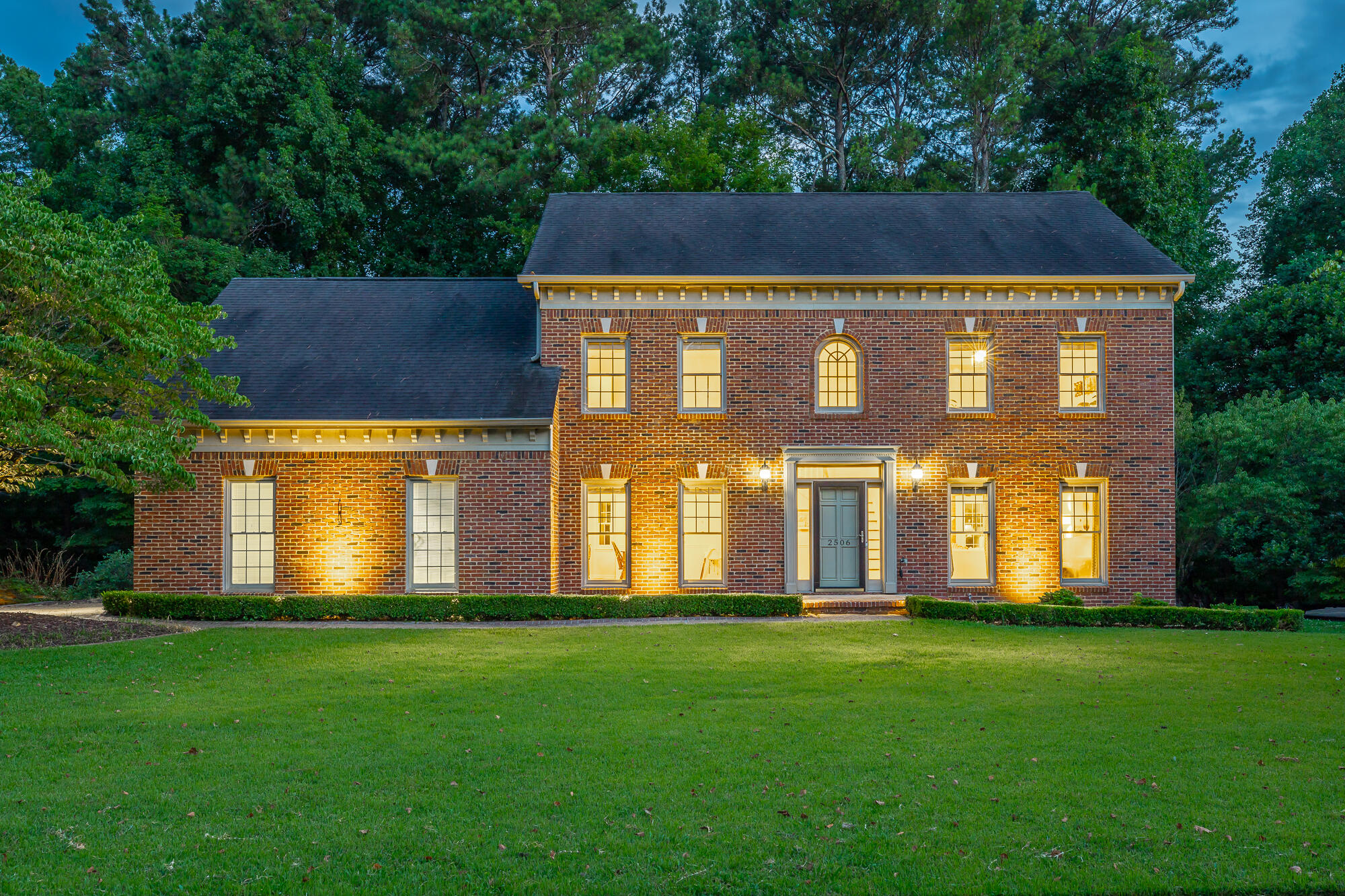 Two-story brick home with glowing windows, a broad lawn, and mature trees at dusk.