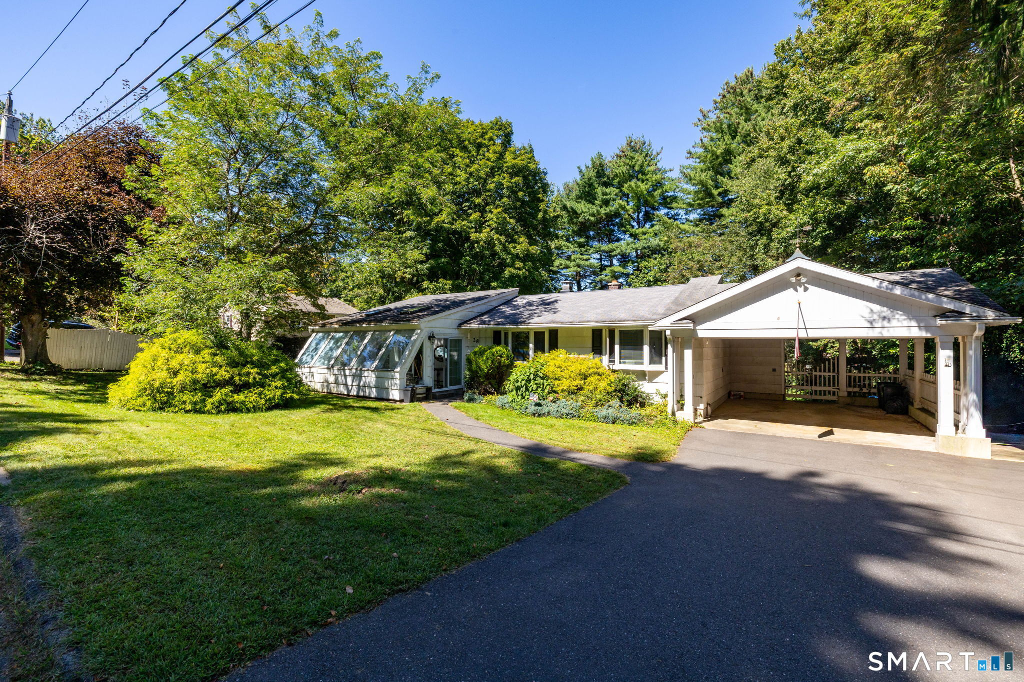 Single-story white house with an enclosed glass sunroom, covered carport, and tree-shaded lawn.