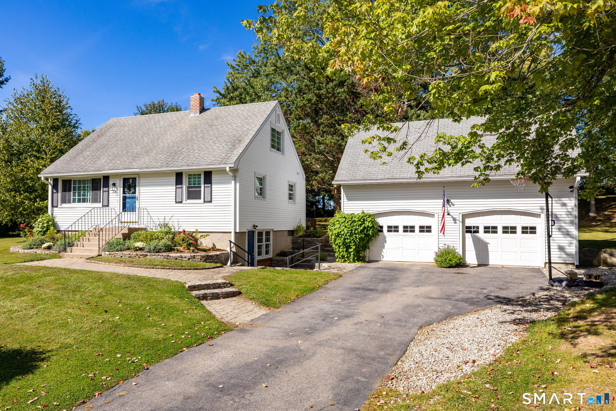 White Cape-style house with blue front door, paved driveway, and two-car garage beneath mature trees.