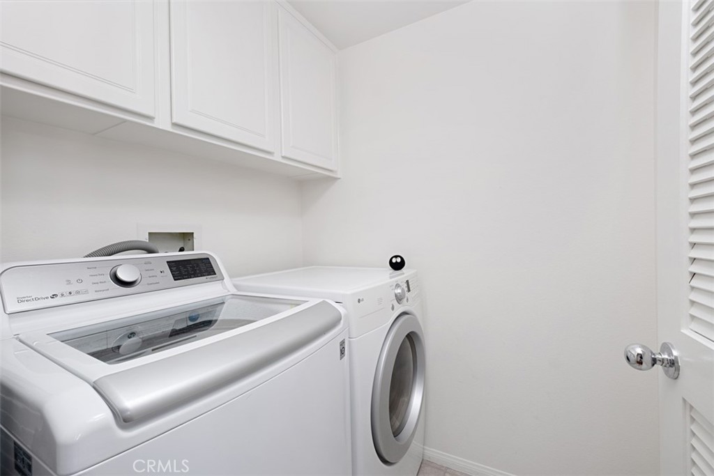Laundry room with white washer and dryer beneath overhead cabinets beside a louvered door.