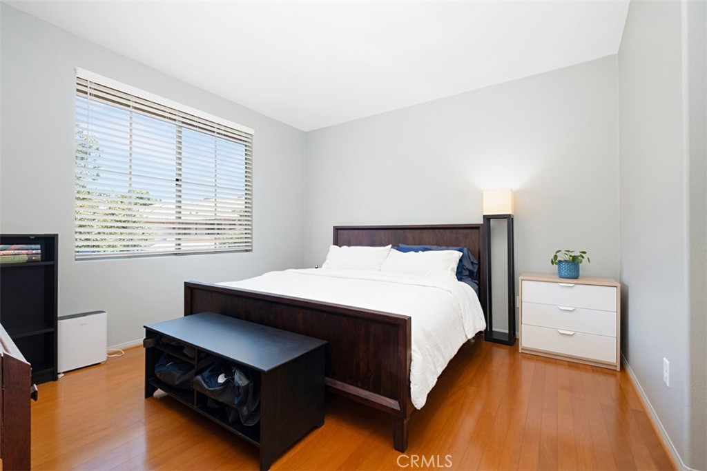 Bedroom with a dark wood bed, white linens, hardwood floors, and a wide window with blinds.