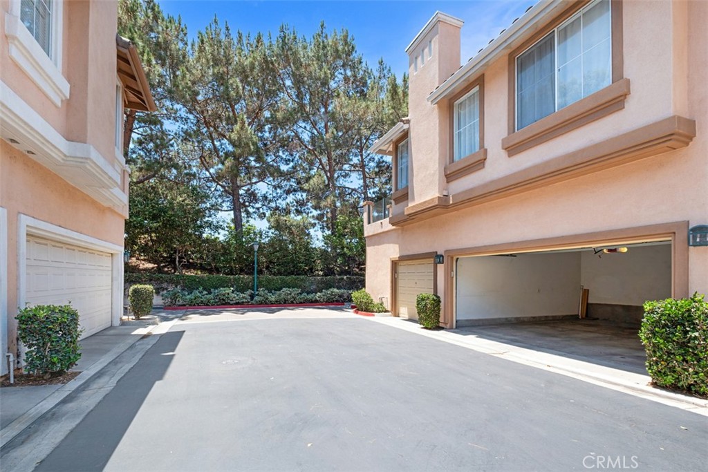 Attached homes with garages facing a shared driveway, bordered by shrubs and mature trees.