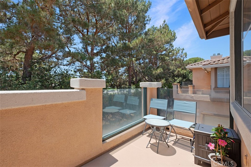 Townhouse balcony with glass railings, patio seating, and tall trees beyond.