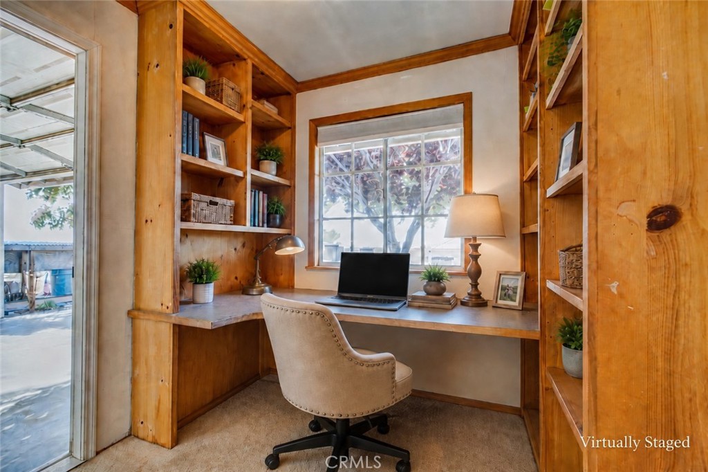 Home office with a built-in wooden desk and shelving, laptop, and upholstered chair by a window.