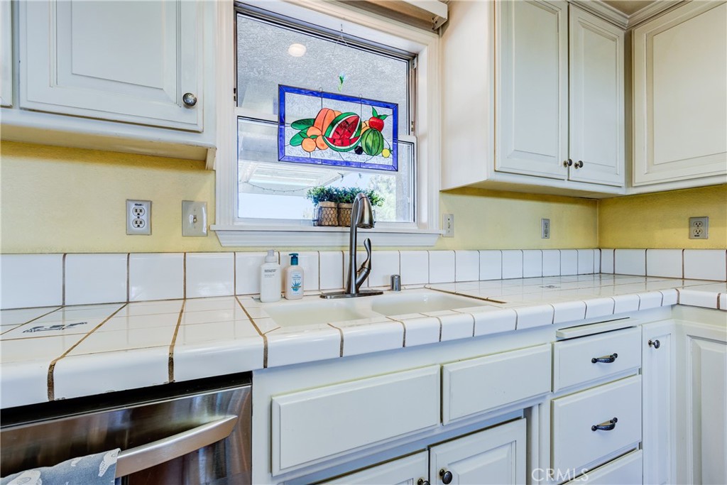 Kitchen sink beneath a window with a colorful fruit stained-glass panel and white tiled counters.
