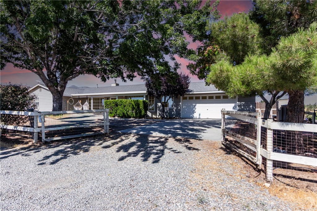 Single-story house with an attached garage, gravel driveway, white fencing, and mature trees.