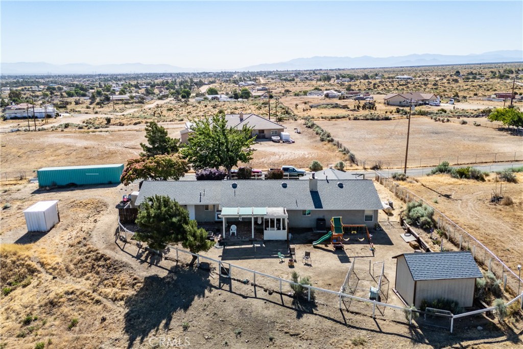 Aerial view of a single-story home with a fenced yard, playset, sheds, and dry open land.