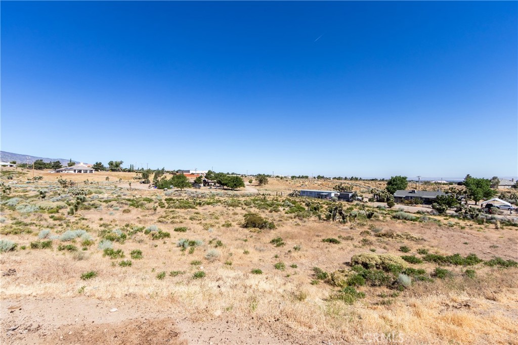 Dry, scrub-covered vacant lot with scattered homes and distant hills beneath a clear blue sky.
