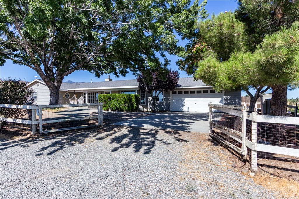 Single-story home with a two-car garage, gravel driveway, open gate, and mature shade trees.