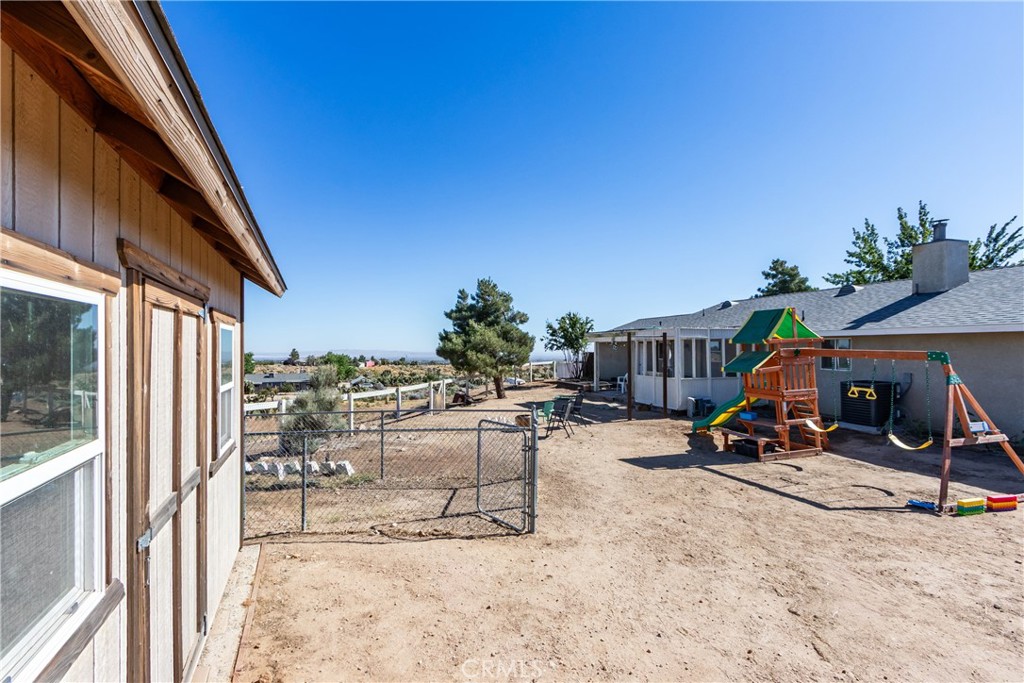 Fenced dirt backyard with a colorful playset and wooden outbuilding beside the house.