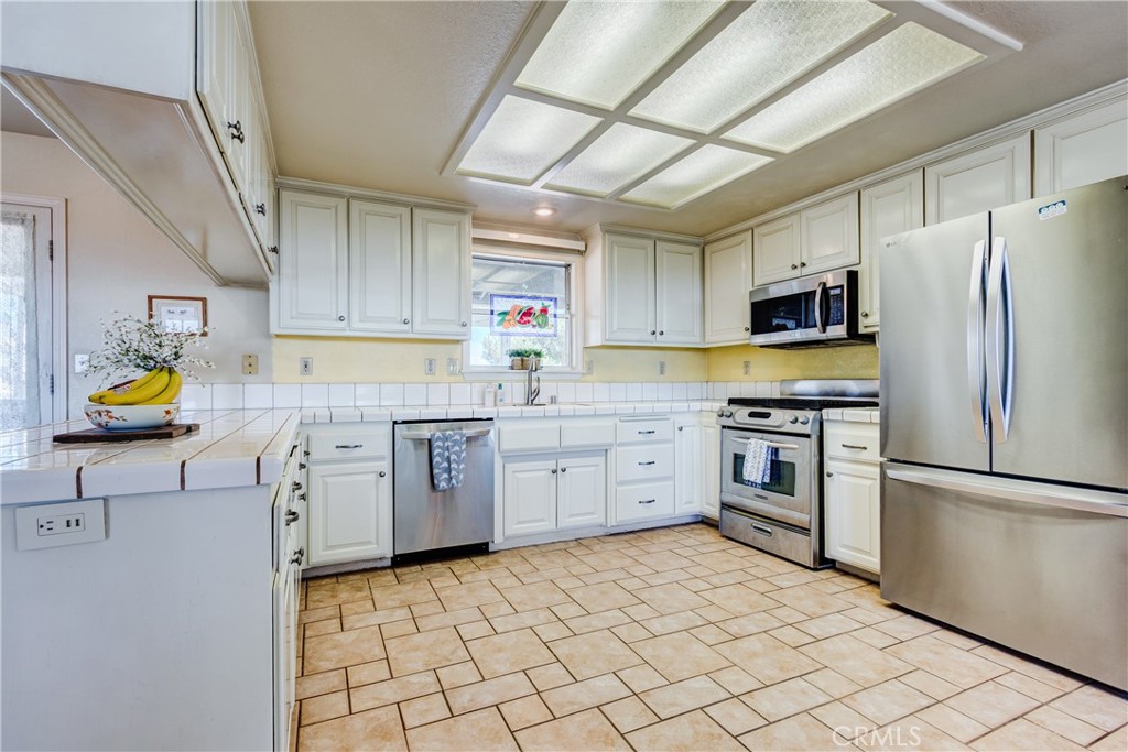 Kitchen with white cabinetry, stainless steel appliances, tiled counters, and tan tile flooring.