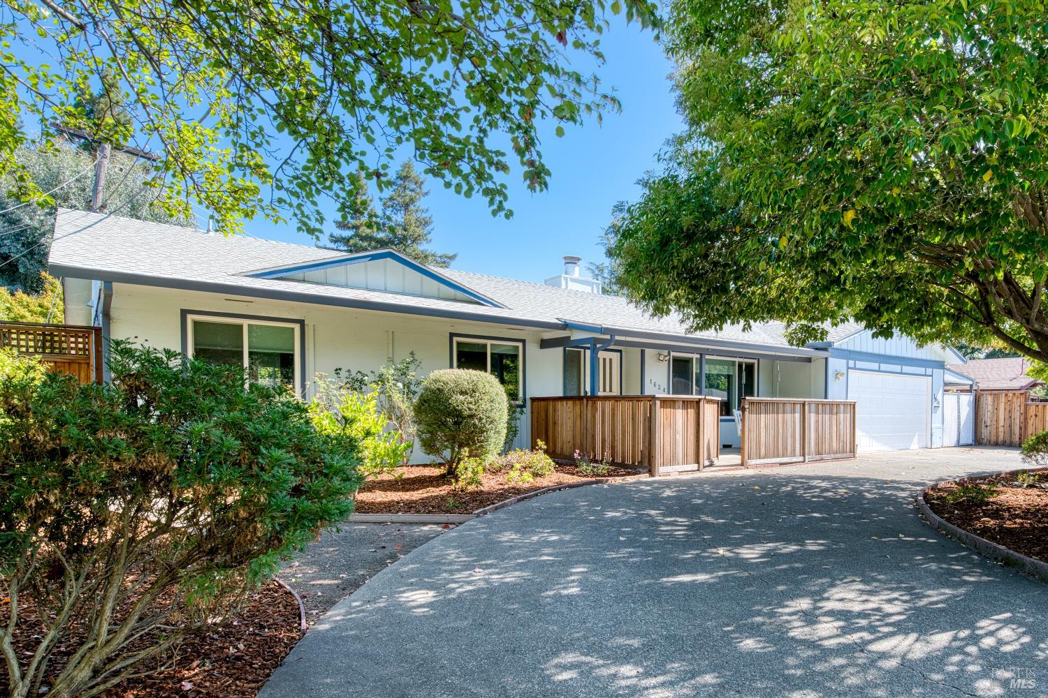 Single-story home with blue trim, a broad driveway, wooden entry fencing, and mature landscaping.