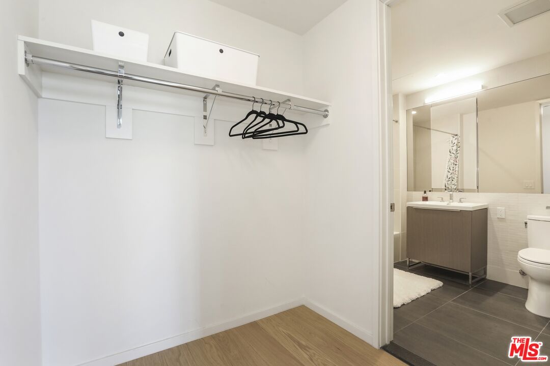 Walk-in closet with a wall shelf and hangers, adjoining a modern bathroom with a vanity and dark tile floor.