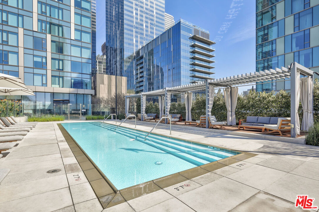 Outdoor pool surrounded by high-rise buildings, with pergola-covered seating and sun loungers.
