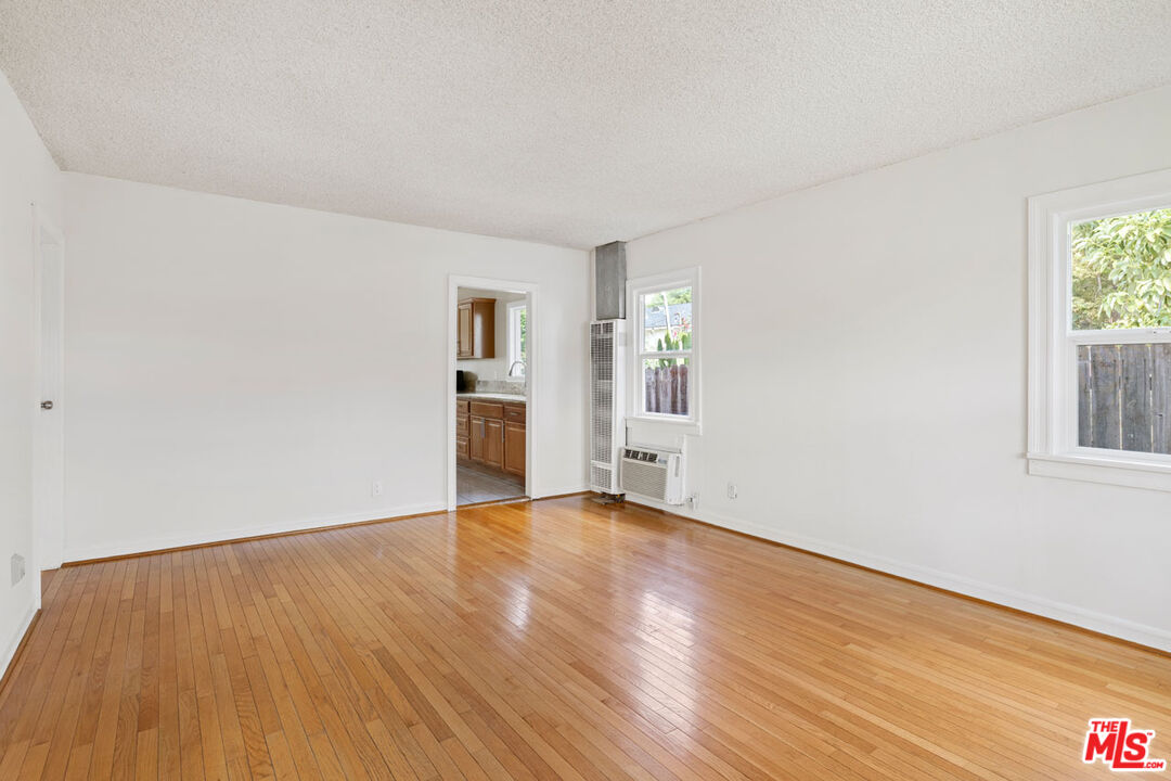 Empty living room with polished hardwood floors, two windows, and a doorway to the kitchen.