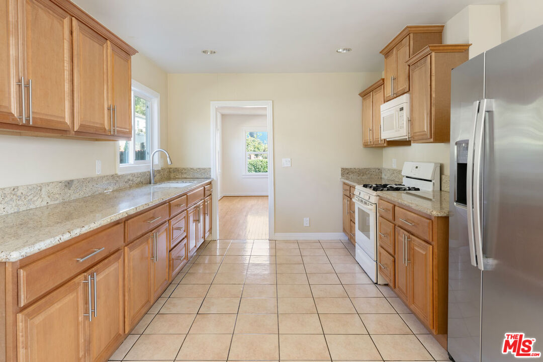Galley kitchen with honey-colored cabinets, stone countertops, stainless steel refrigerator, and beige tile floor.