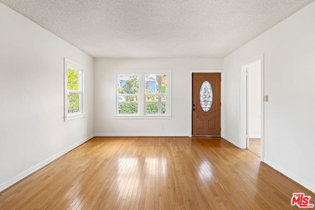 Empty front room with polished hardwood floors, two windows, and a wooden entry door with decorative glass.