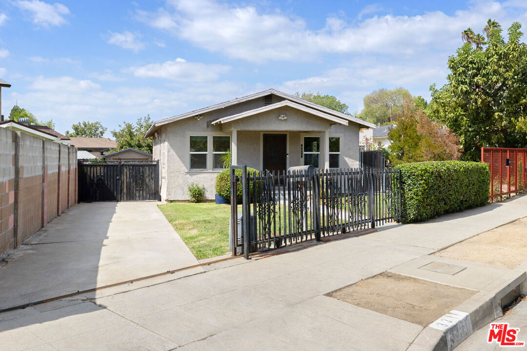 Single-story stucco house behind black metal gates, with a driveway and trimmed hedge.