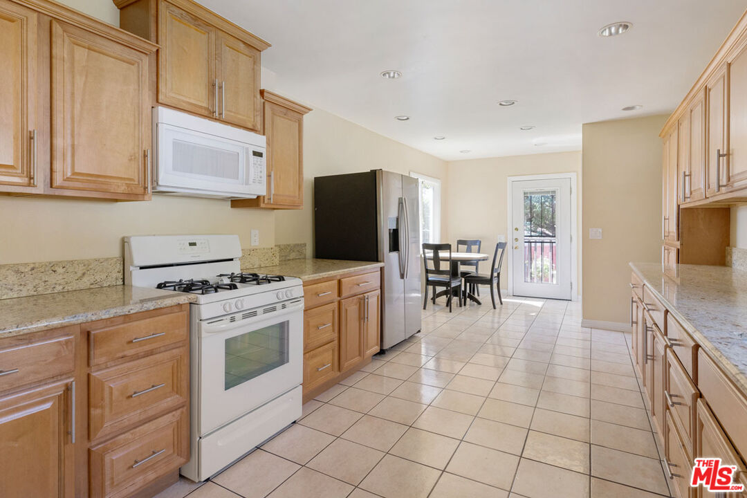 Kitchen with light wood cabinetry, stone countertops, stainless steel refrigerator, and dining area by a glass door.