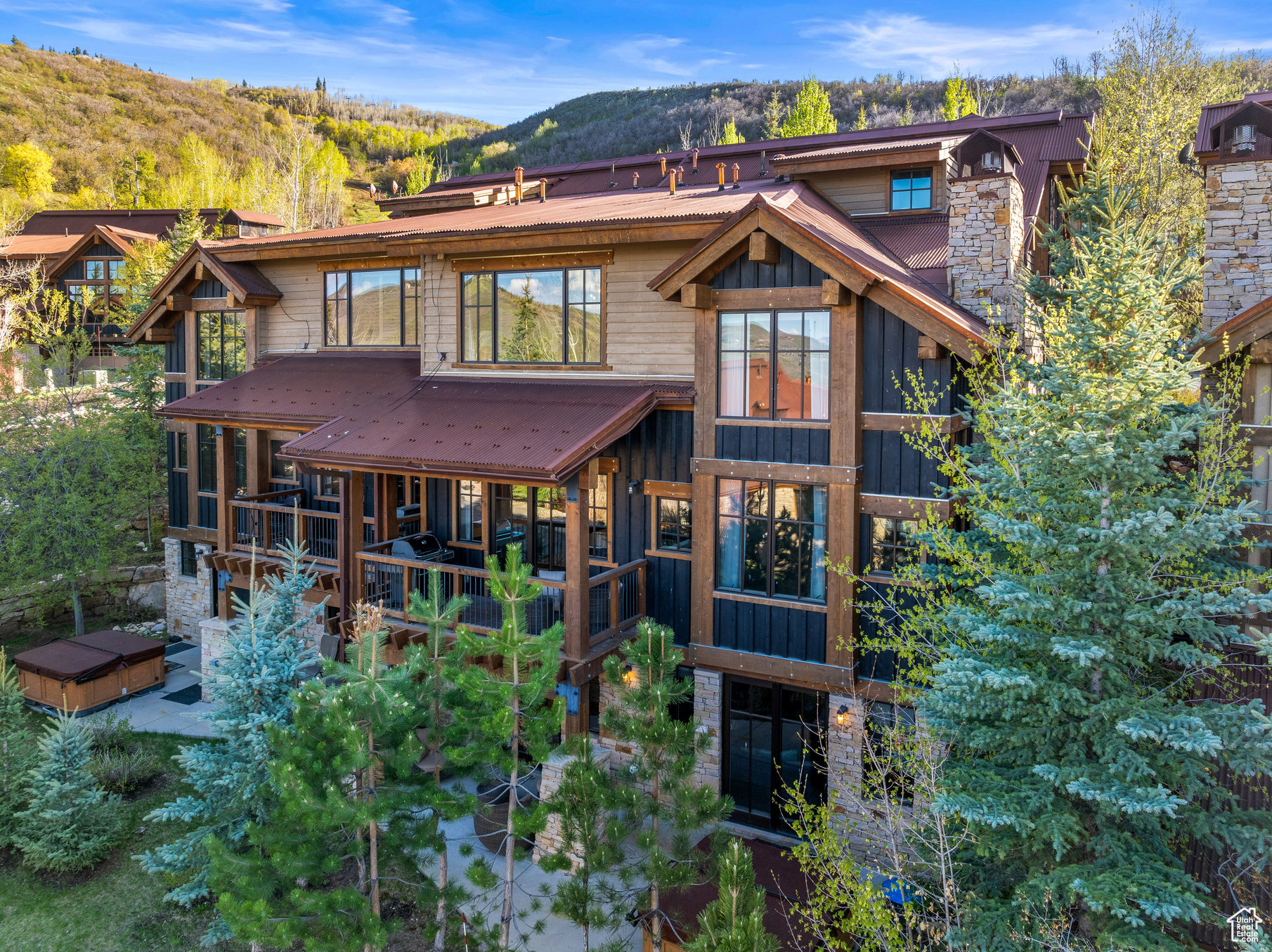 Mountain-style home with timber framing, stone accents, covered decks, and a hot tub among evergreen trees.