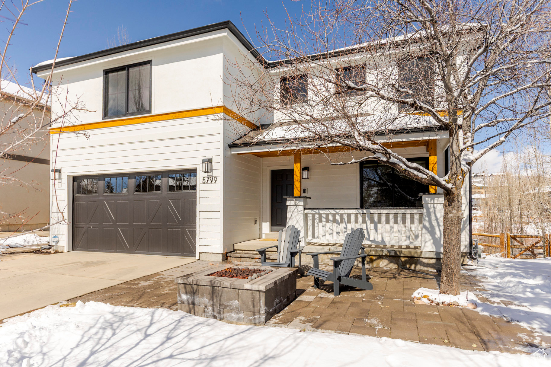 Modern white two-story home with a dark garage, covered porch, patio chairs, and snow-covered yard.