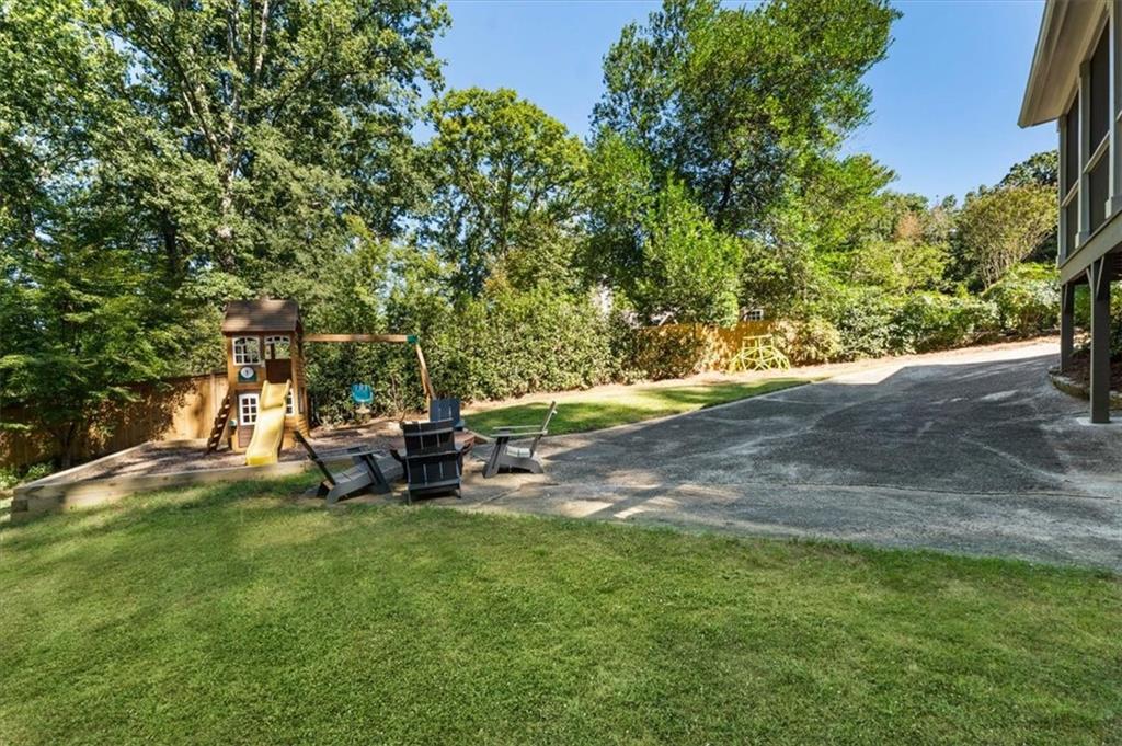 Backyard lawn with a children's playset, yellow slide, Adirondack chairs, and sloping driveway.