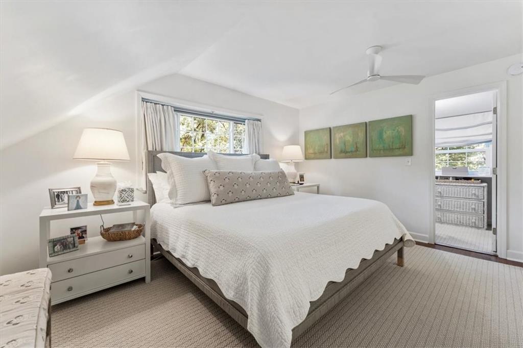 Bright bedroom with a large white bed, sloped ceiling, ceiling fan, and adjoining room visible through an open doorway.