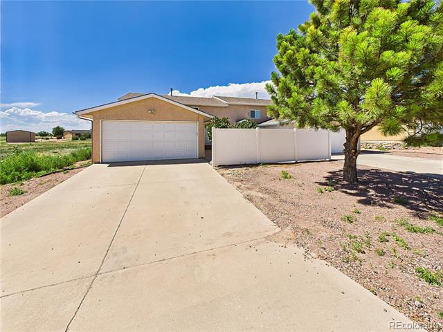 Tan detached garage with a wide concrete driveway, white fence, and large evergreen tree.