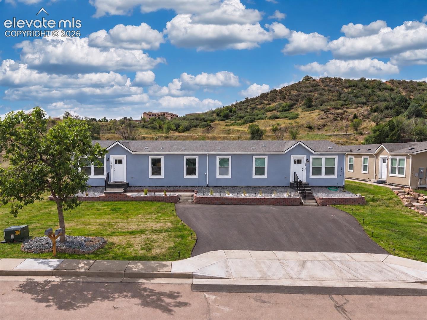 Row of single-story homes with paved driveway and a scrub-covered hillside behind them.
