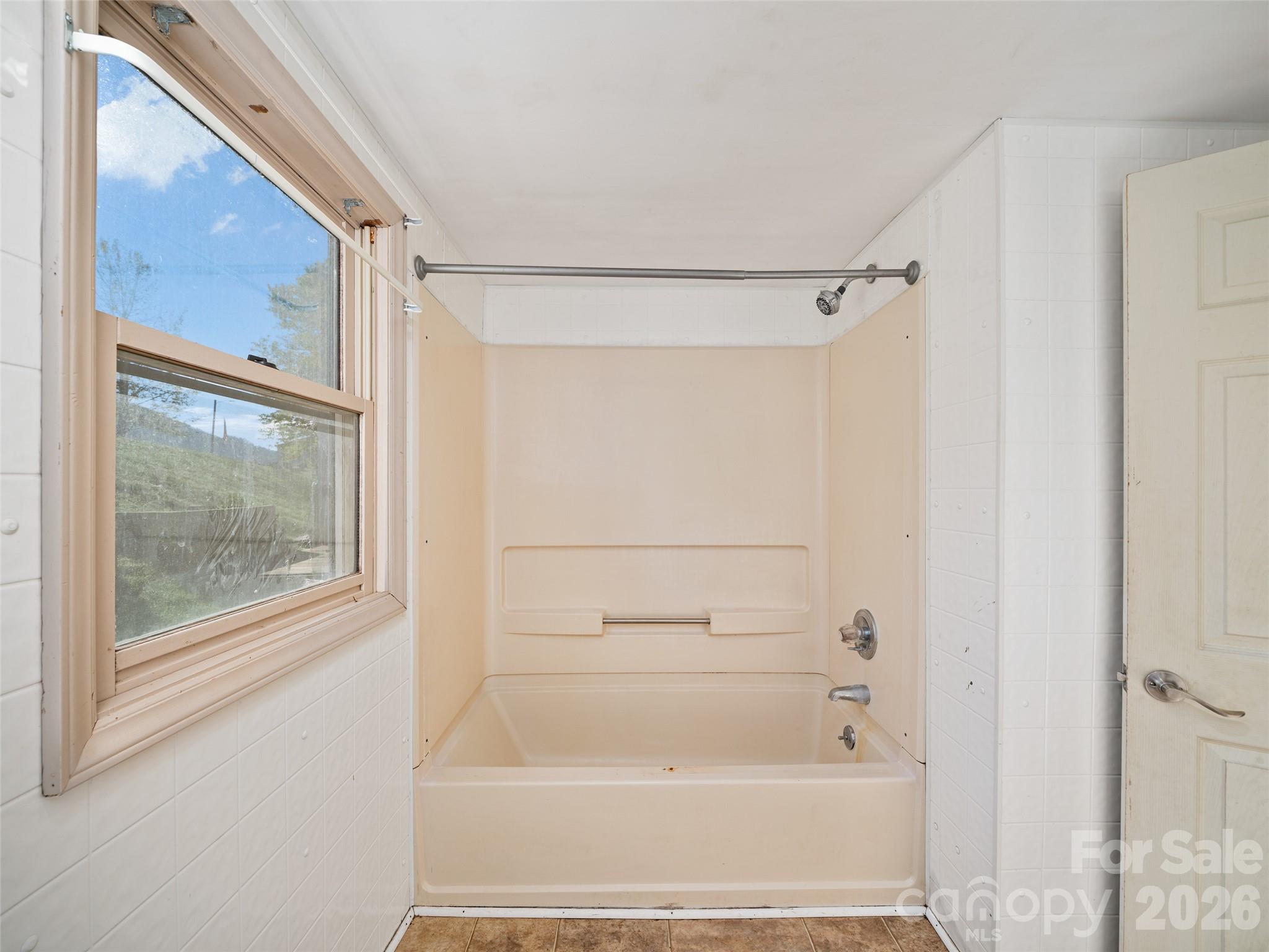 Bathroom tub and shower beneath a window, with white tile and an open door.