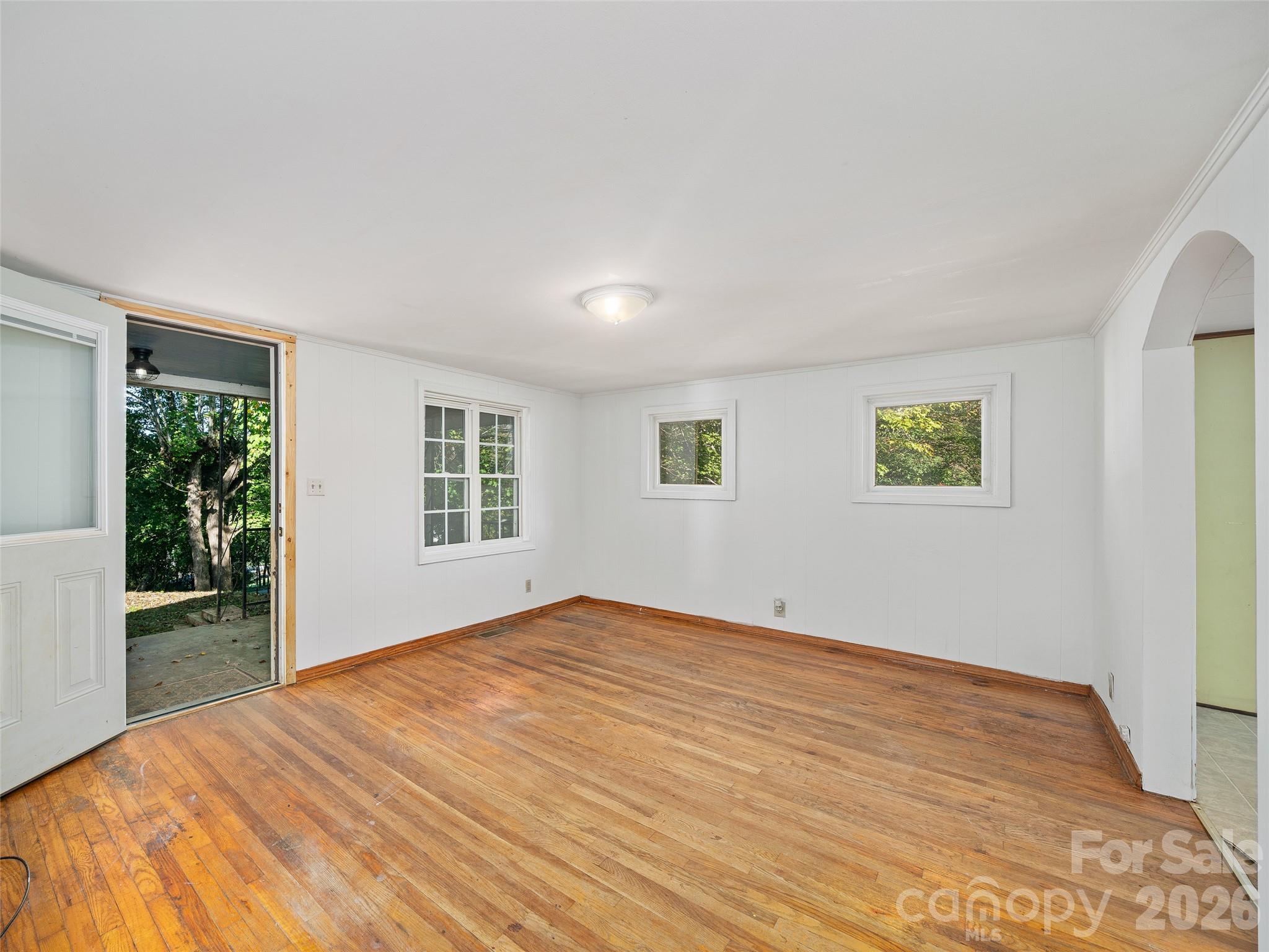 Empty living room with hardwood floors, white walls, and an open door to a wooded yard.