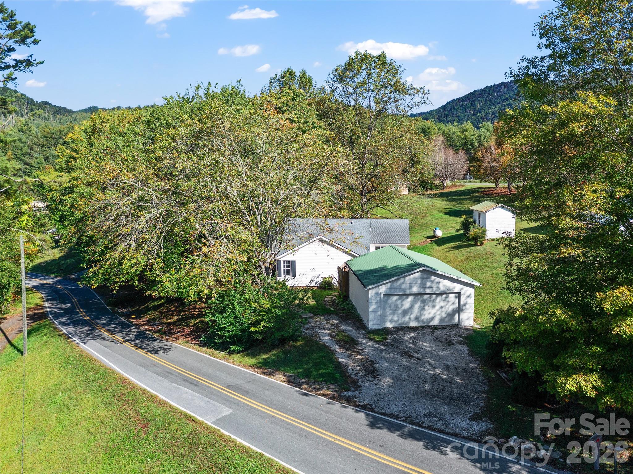 Aerial view of a white house and detached garage beside a winding road, with wooded mountain views.