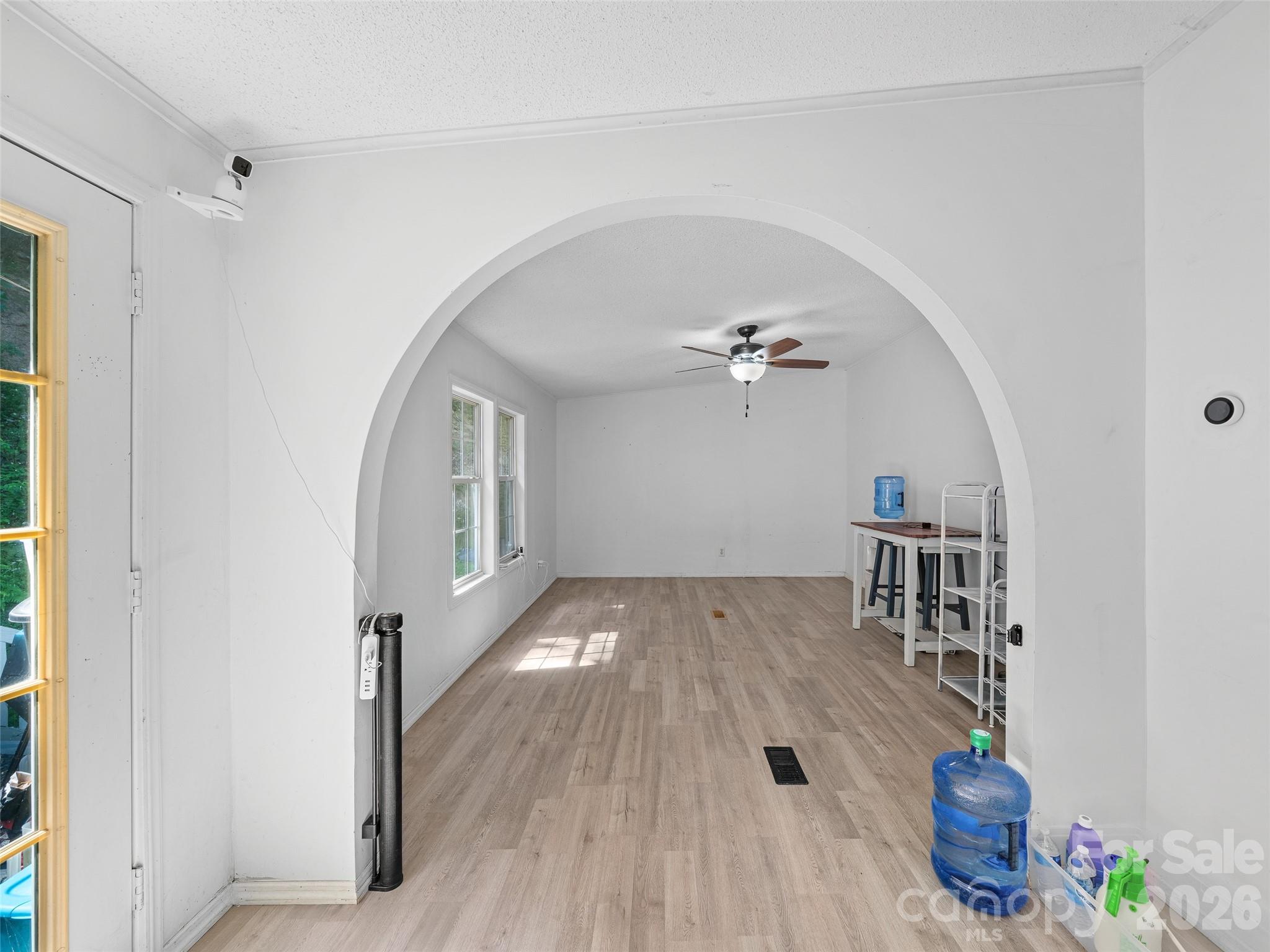 Bright, mostly empty room with a large arched opening, wood-look floors, two windows, and a ceiling fan.