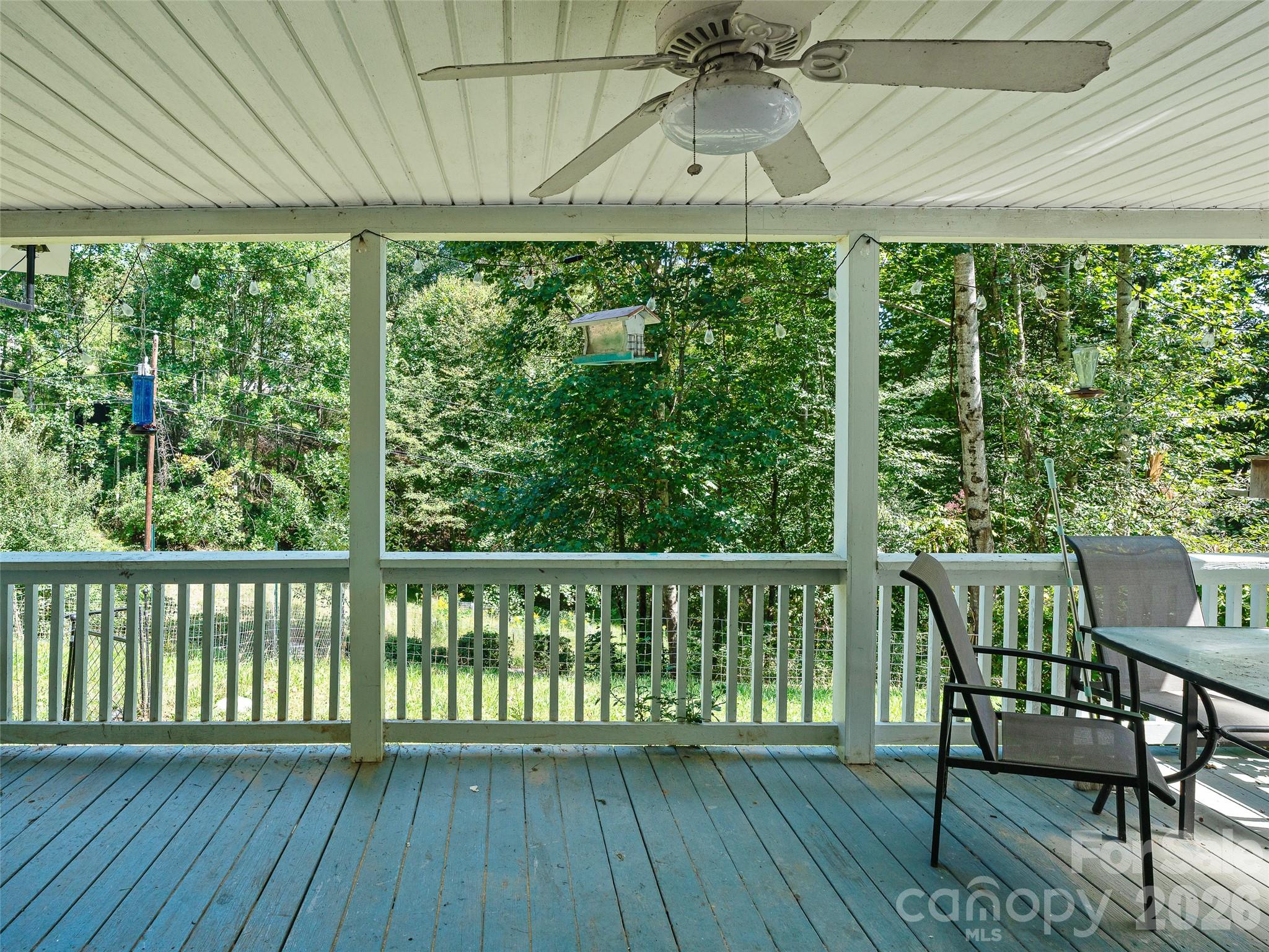 Covered porch with blue floorboards, white railings, and chairs overlooking wooded surroundings.