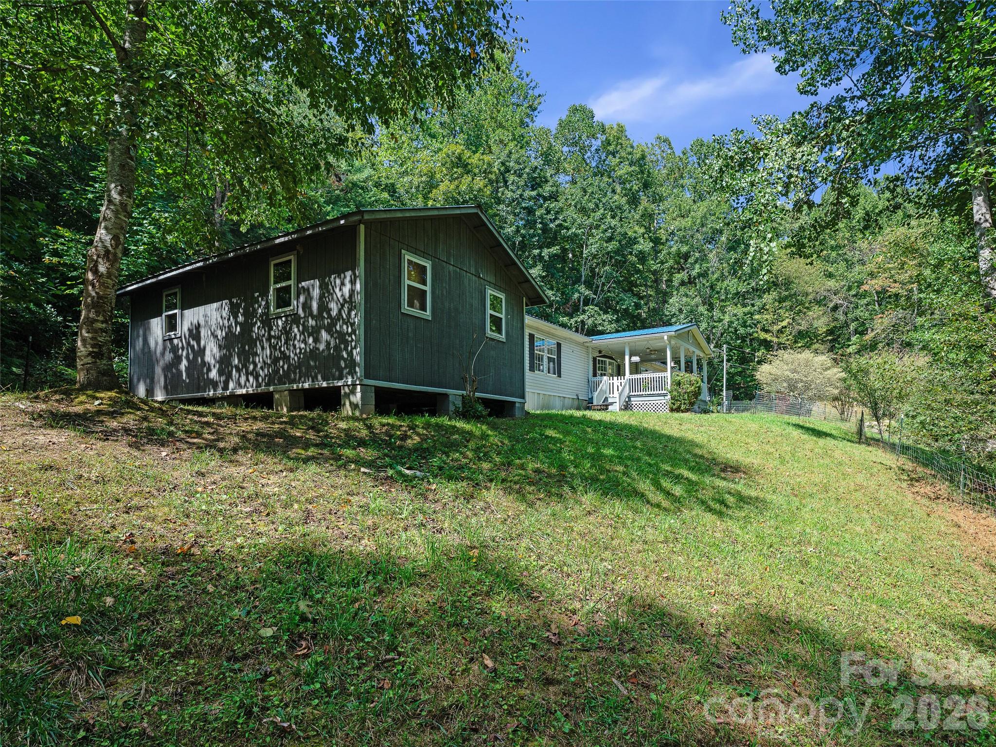 Dark-sided home with a white covered porch, set on a grassy slope among trees.