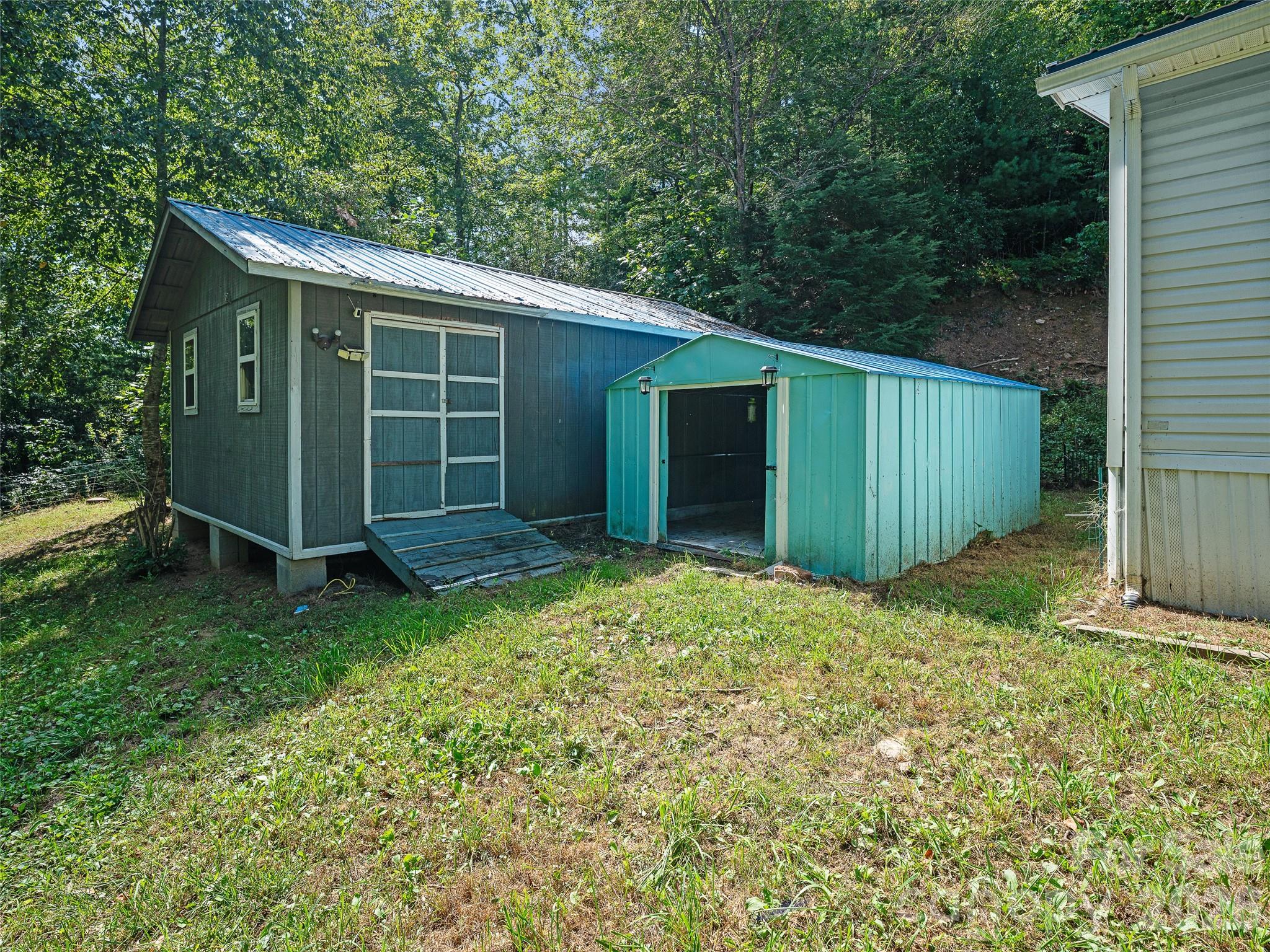 Two sheds beside a home, including a large gray shed with a wooden ramp and an open turquoise shed.
