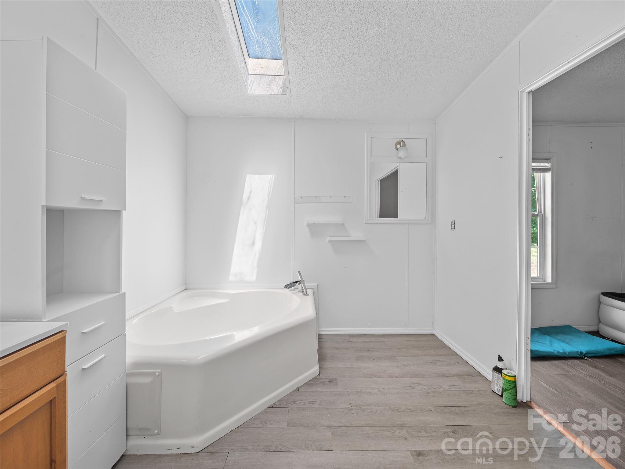 Bathroom with a large corner tub, built-in cabinetry, skylight, and gray wood-look flooring.