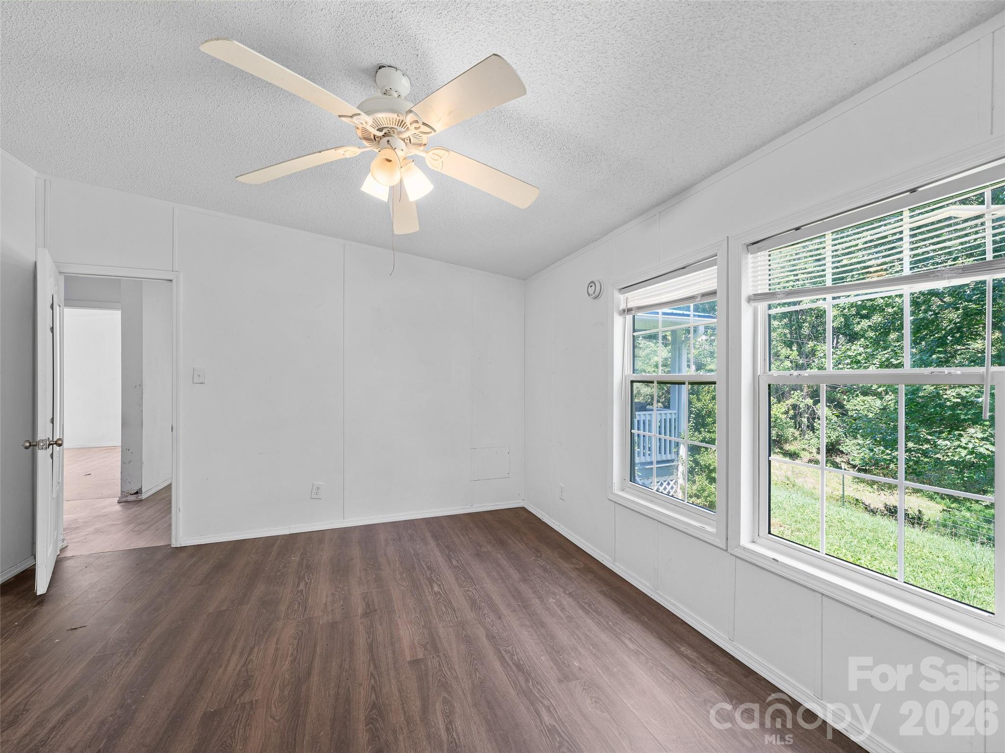 Empty room with dark flooring, a ceiling fan, and two large windows overlooking a green yard.