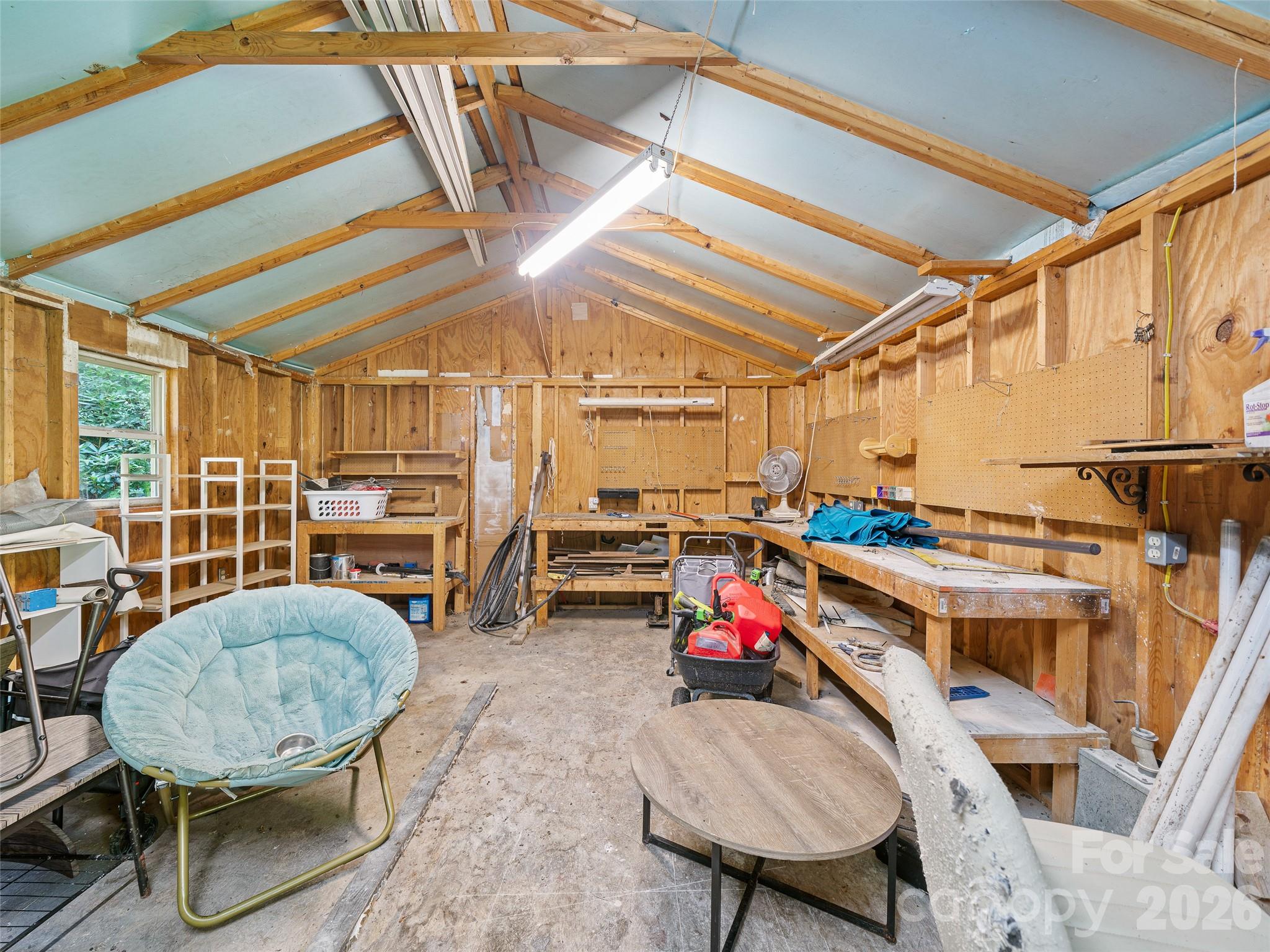 Workshop with exposed rafters, wall-mounted pegboards, workbenches, tools, and a pale blue lounge chair.