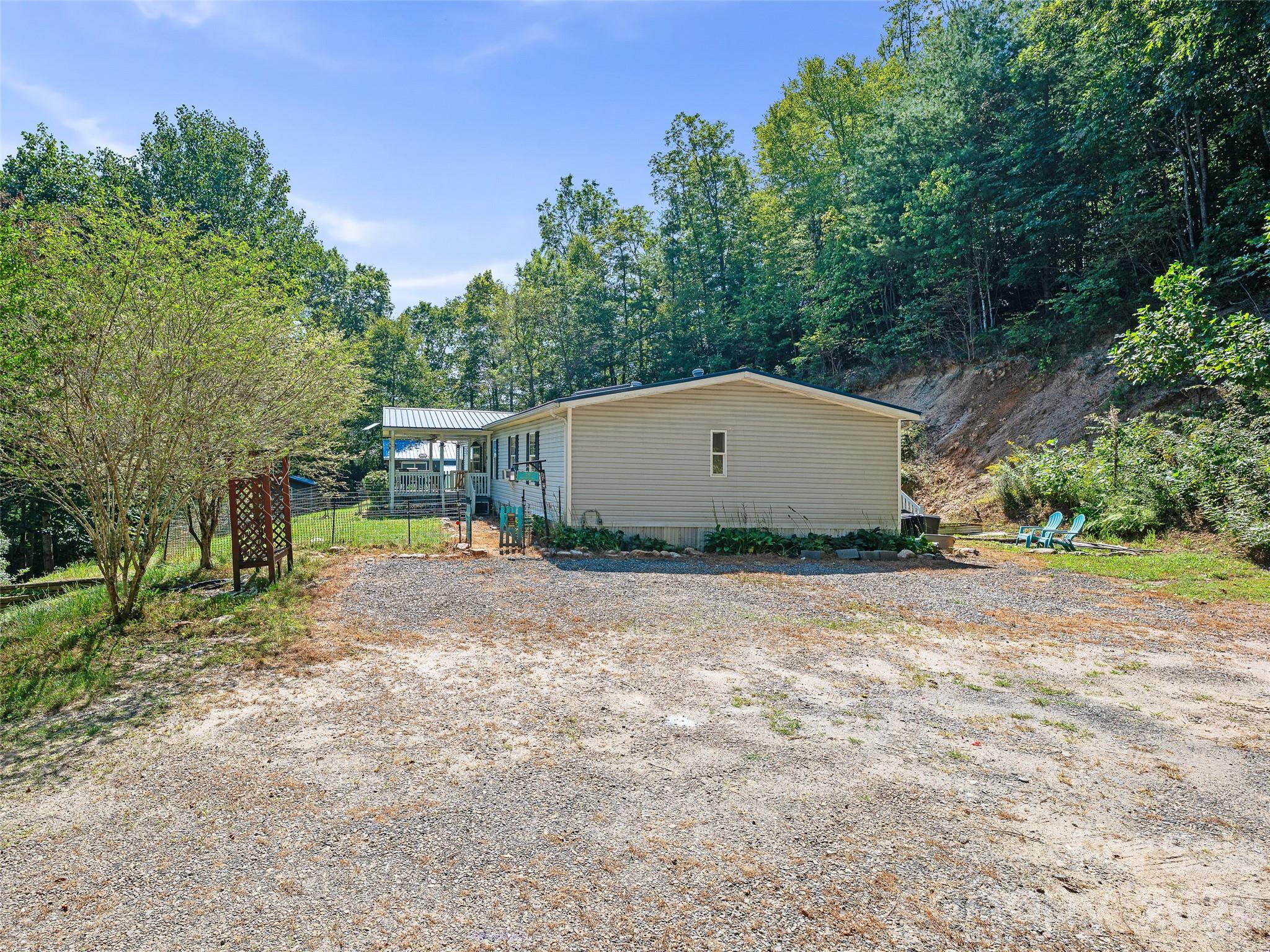 Beige single-story home with a covered porch, broad gravel driveway, and wooded surroundings.