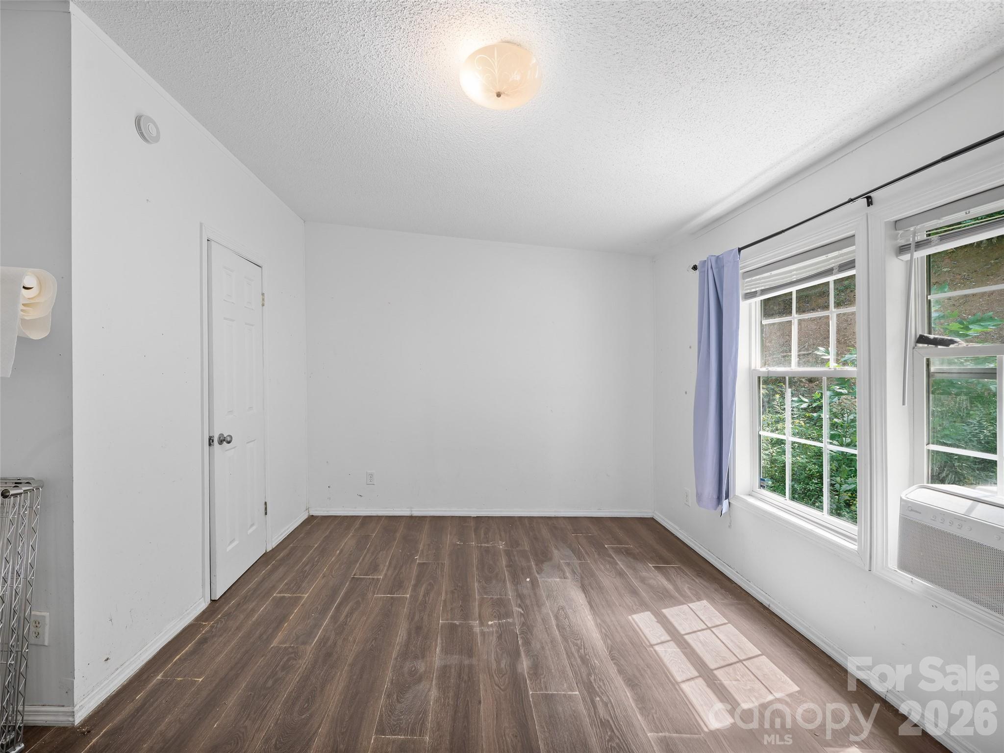 Empty room with dark wood-look flooring, two windows, a lavender curtain, and a window air conditioner.