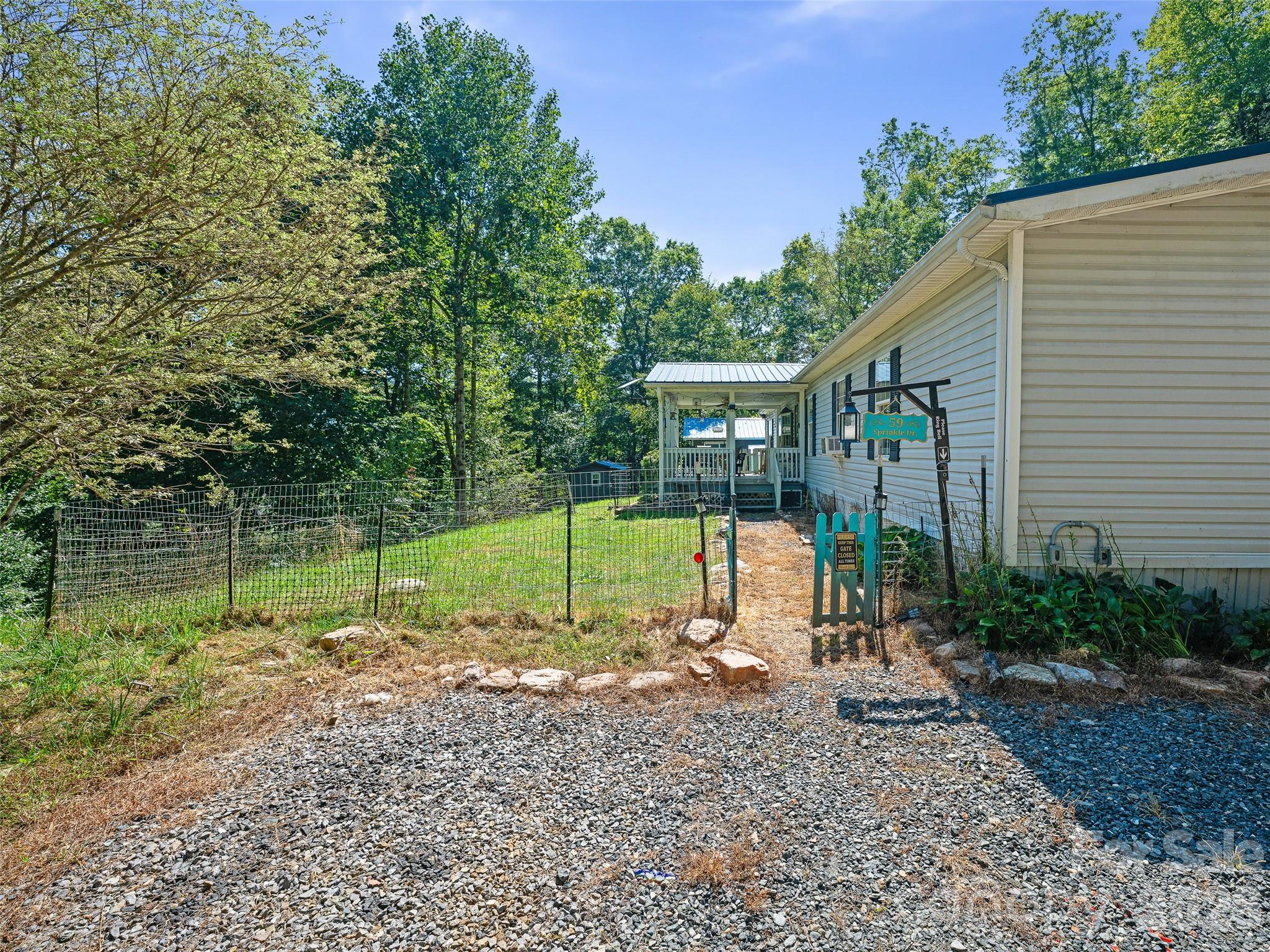 Home exterior with a covered porch, fenced yard, and gravel driveway beneath leafy trees.