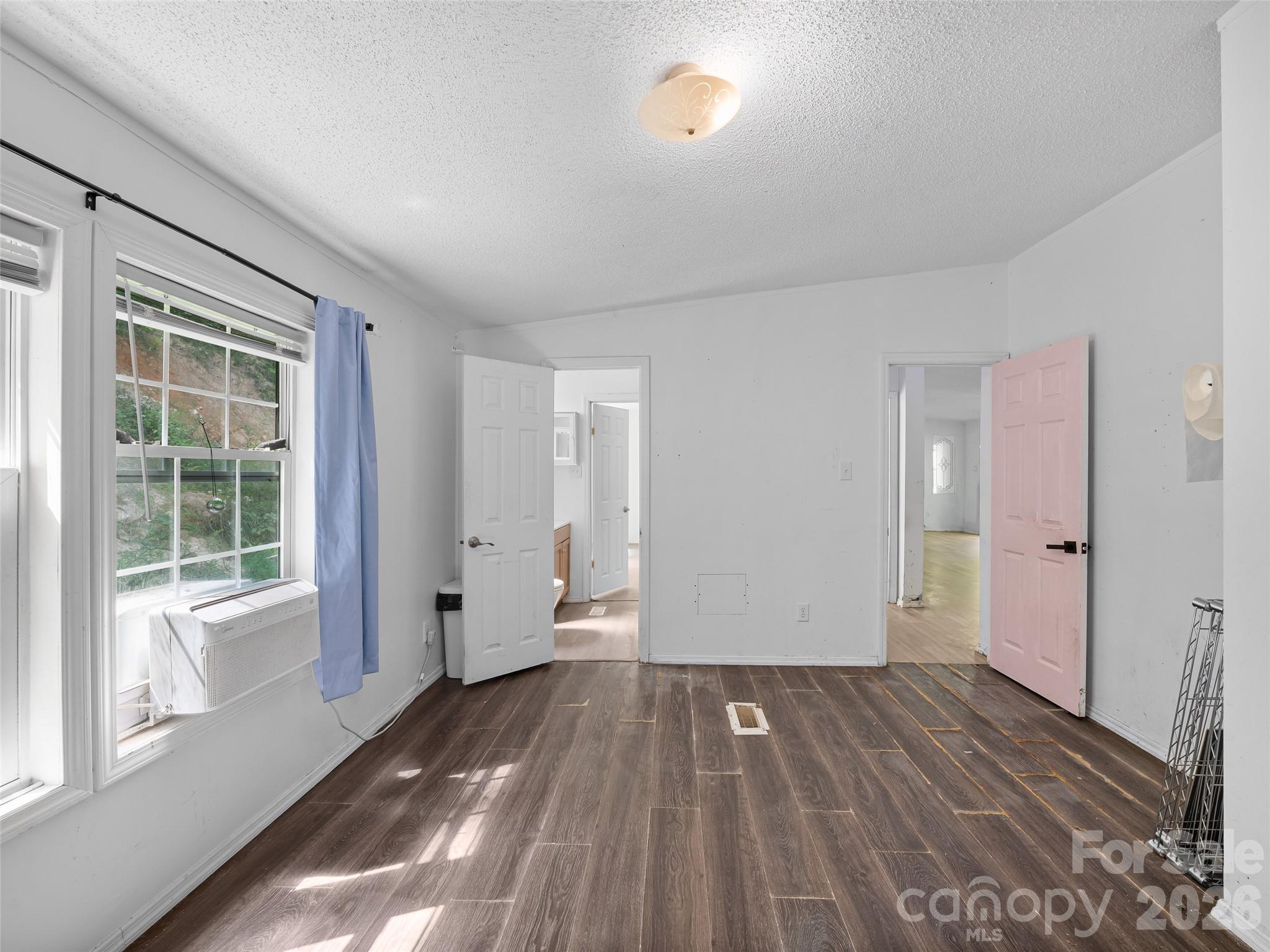 Empty bedroom with dark wood-look flooring, window air conditioner, blue curtain, and open pink door.